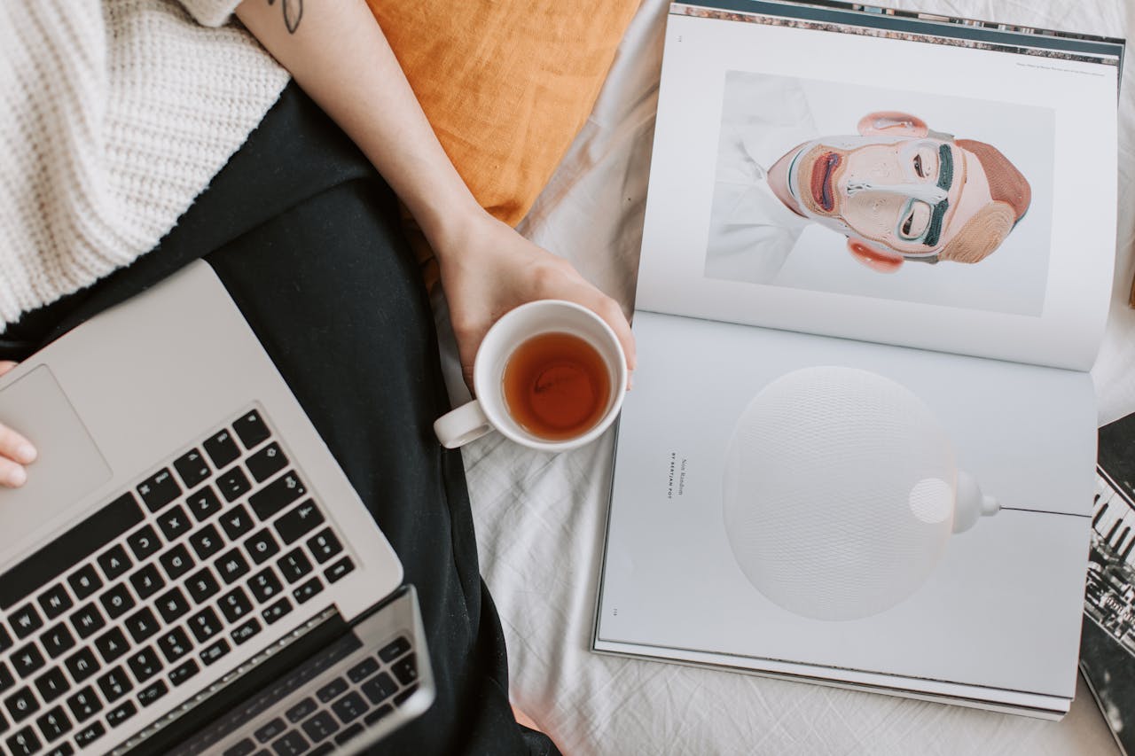 about-us Woman relaxing with tea, browsing laptop, and reading magazine in a cozy home setting.